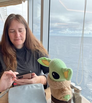 a woman sitting at a table looking at phone with a little green companion