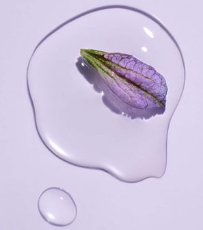 A close-up of a purple flower petal resting on a clear liquid, symbolizing the gentle and hydrating 