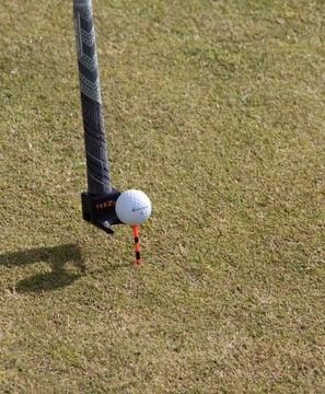 A golf club with a 'TeeZUP' attachment holding an orange tee and a golf ball, set up on a grassy course, ready for a shot