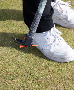 Person picking up a golf tee of the grass using the TeeZUP tool on grass, with a club and white sneakers visible.