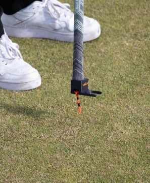 A golfer uses the 'TeeZUP' tool, attached to the end of a golf club, to pick up a orange tee in place on the grass