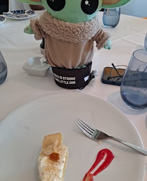 a little green companion standing on table in front of a plate with a slice of cheesecake on it