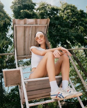 Model lounges on an umpire chair in retro sunglasses and a white baby tee, framed by tropical outdoor light.
