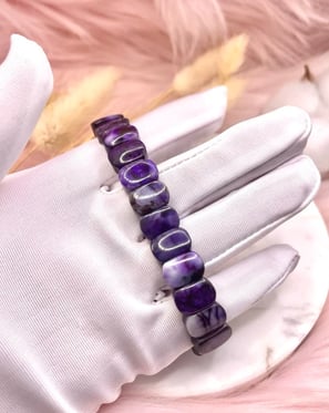 A gloved hand holding a deep purple beaded bracelet against a soft, neutral-toned background.