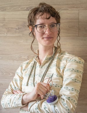 Une personne souriante sur un fond de bois. Elle a des outils de couture en main