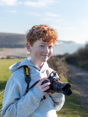 a profile photo of Drew Walker, smiling holding a Nikon D3300 with a background of Cuckmere Haven and the seven sisters.