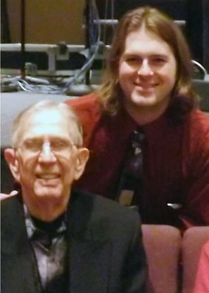 Photograph: a 30-ish white person with shoulder-length blond hair and a tie leans over a squinting, 90-ish man in a dark suit