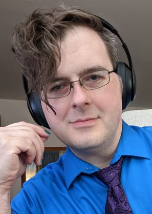 photo of GJ, white adult, with asymetrical wavy hair, glasses, chunky headphones, graying temples, nailpolish, & paisley tie