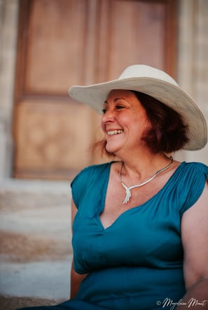 Portrait de Flo Salort assise sur des marches, sourire et chapeau, incarnant liberté et féminité