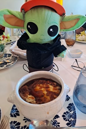 Little green companion wearing a black suit standing in front of bowl of French onion soup 