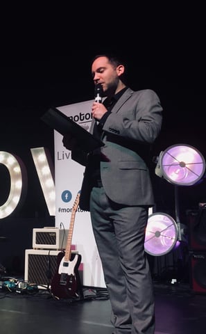 Alex hosting an awards function in a grey suit holding a clipboard