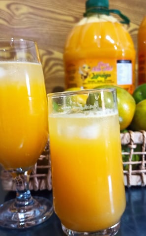 Taças de suco de laranja sobrepostas em uma mesa. Ao fundo, garrafas de suco de laranja.