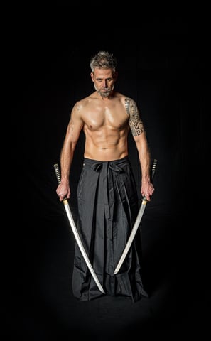Ein muskulöser, tätowierter Mann mittleren Alters steht mit zwei Katanas auf einem schwarzen Hintergrund. Er trägt einen trad