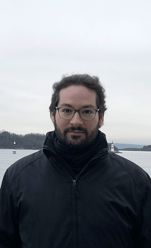 Photo of a man wearing glasses, with a slight smile. Behind him is the sea, a lighthouse and a forest. It is cloudy.