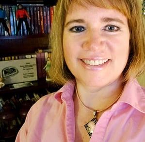 Susan Gable wearing a pink shirt, smiling, posing in front of her bookshelf.