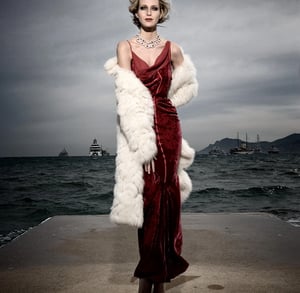 Model wearing evening dress and coat standing on a dock with the ocean and ships in the background