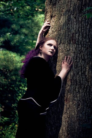 a woman with purple hair in a black dress in the forest with sunlight on her face