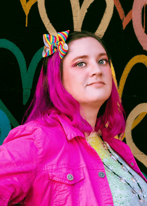 Erika Gritton stands in front of a wall of painted hearts. Her hair is pink with a rainbow bow in it
