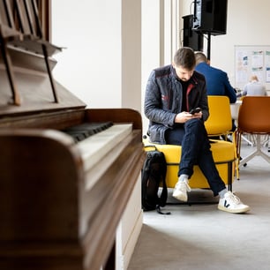 Une homme en veste noire assis sur un fauteuil jaune et regardant son smartphone