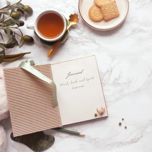 Marble table top with journal, olives and cup of tea with biscuits on a plate.