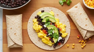The image shows a breakfast wrap with scrambled eggs, black beans, avocado, cilantro, and tomato, ready to be rolled up.
