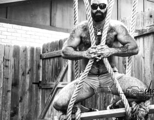 hairy, shirtless bearded man in sunglasses and cutoff shorts hugging ropes