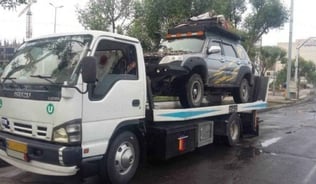 Caminhão branco, tipo plataforma, carregando um veículo de trilha ou rally, o asfalto está molhado.