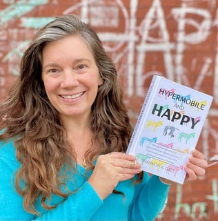 pretty woman with long brown hair in a turquoise sweater holding a book in front of a brick wall