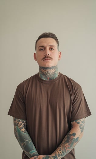 Portrait d’un homme tatoué, debout devant un fond neutre. Il a une moustache, un piercing au nez,
