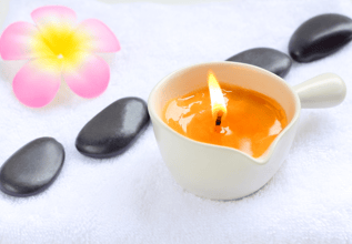 a massage candle in a cup container over a white cloth table with mindfulness stone and a flower