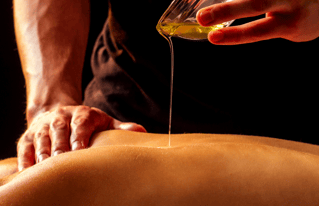 A hand pours oil from onto the back of a lying woman, another hand rests on the woman's shoulder