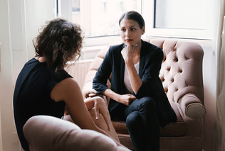 Life coach sitting on a sofa chair coaching a client sitting on a coach opposite of her.