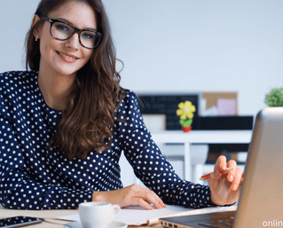 a woman in glasses and a polka dot shirt is smiling and holding a pen with a laptop on the desk