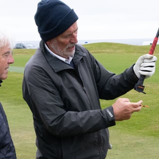 Two men on a golf course, one holding a golf club with a TeeZUP tool attached demonstrating the use of a tee to the other man