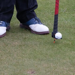 A golfer wearing two-tone shoes, standing near a golf ball balanced on an orange tee using the 'TeeZUP' tool