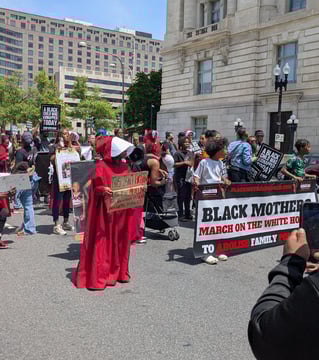 Front of the marching line. People are holding a banner and a woman is dressed like a Handmaid. 