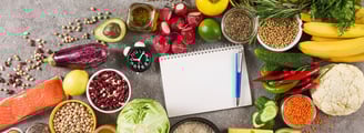 A colourful array of healthy food, vegetables and grains, with a notepad and pen and a clock in the middle