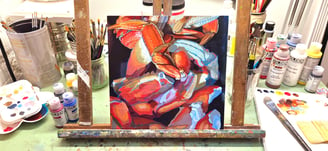 An in-progress painting sits on an easel on a desk cluttered with paints, brushes and palettes.