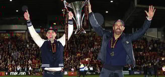 "Ryan Reynolds and Rob McElhenney celebrating on the football pitch, holding the Vanarama National L