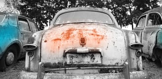 Three abandoned vintage cars in a field – a turquoise sedan flanked by rusty companions beneath shadowy trees.