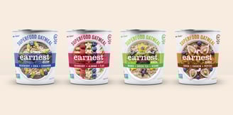 Four 3D mockups of Earnest Eats oatmeal cups side by side on a beige background.