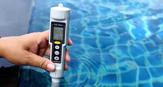 Hand holding a digital salt meter to test pool water quality with reflection of blue tiles.