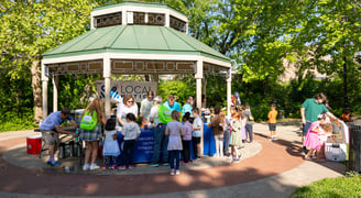 A bunch of children with a few adults approaching a table of adults in a park