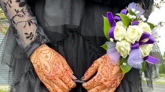 Close up of two hands with henna on them, holding a purse strap, wearing a black dress & corsage
