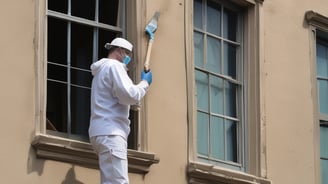 People are working on the interior renovation of a room that appears to be under construction. A worker on a scaffold is painting the ceiling, while others are seated or discussing the progress. The room has smooth, gray walls with arched recesses and a modern design vibe. There are tools and materials scattered around, including a ladder, paint cans, and some rocks or construction debris on the floor. Overhead, stylish pendant lights provide illumination as natural light streams in from windows.
