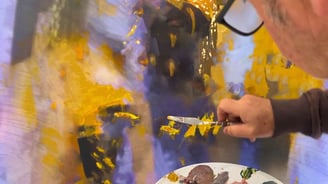 Martin Osner painting with a palette knife on an abstract canvas in gold, violet, and charcoal tones.