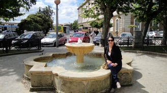 Foto da autora sentada numa fonte na praça próxima ao porto de Cádiz.
