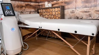 a white massage bad with a white lether top and a white HIFEM Machine on the left side. Wooden wall
