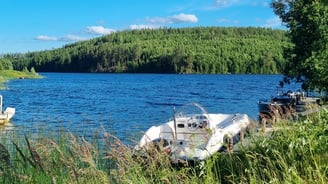 The view of the summerhouse shoreline with a boat nearby, surrounded by water and forest.