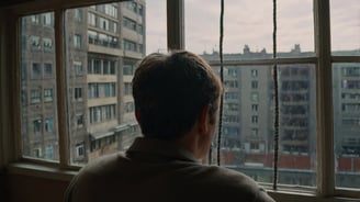 An image of a man with brown hair looking out the window of their apartment onto a grey city